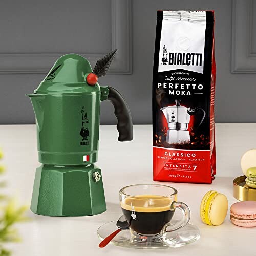 Green Bialetti Moka pot with coffee bag and cup of espresso on table.