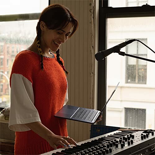 Woman using a laptop while playing a keyboard near a microphone.